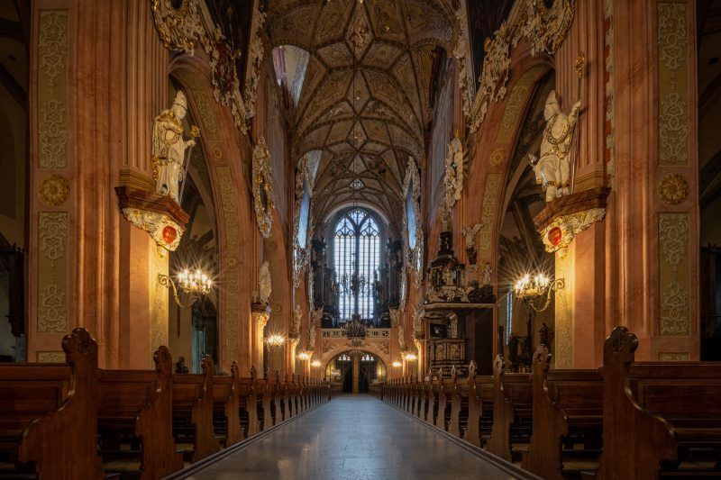Przestronna nawa główna, pełna światła wpadającego przez witraże, z bogato zdobionymi sklepieniami i wysokimi filarami, które prowadzą wzrok ku wielkim ołtarzom i rzeźbom.