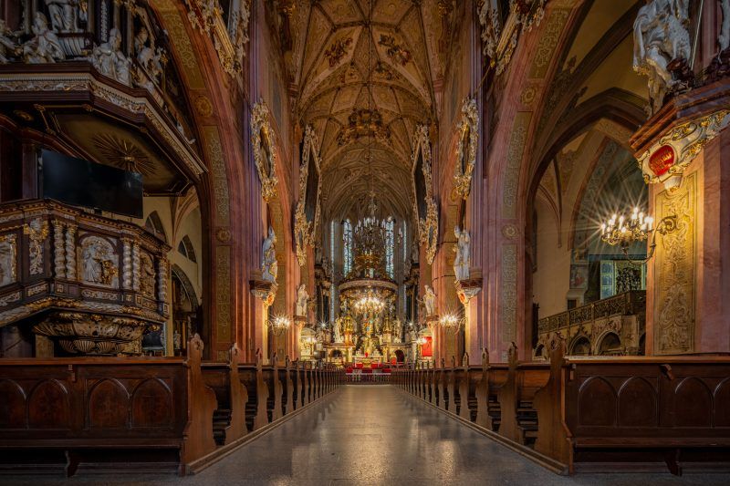 Przestronna nawa główna, pełna światła wpadającego przez witraże, z bogato zdobionymi sklepieniami i wysokimi filarami, które prowadzą wzrok ku wielkim ołtarzom i rzeźbom.