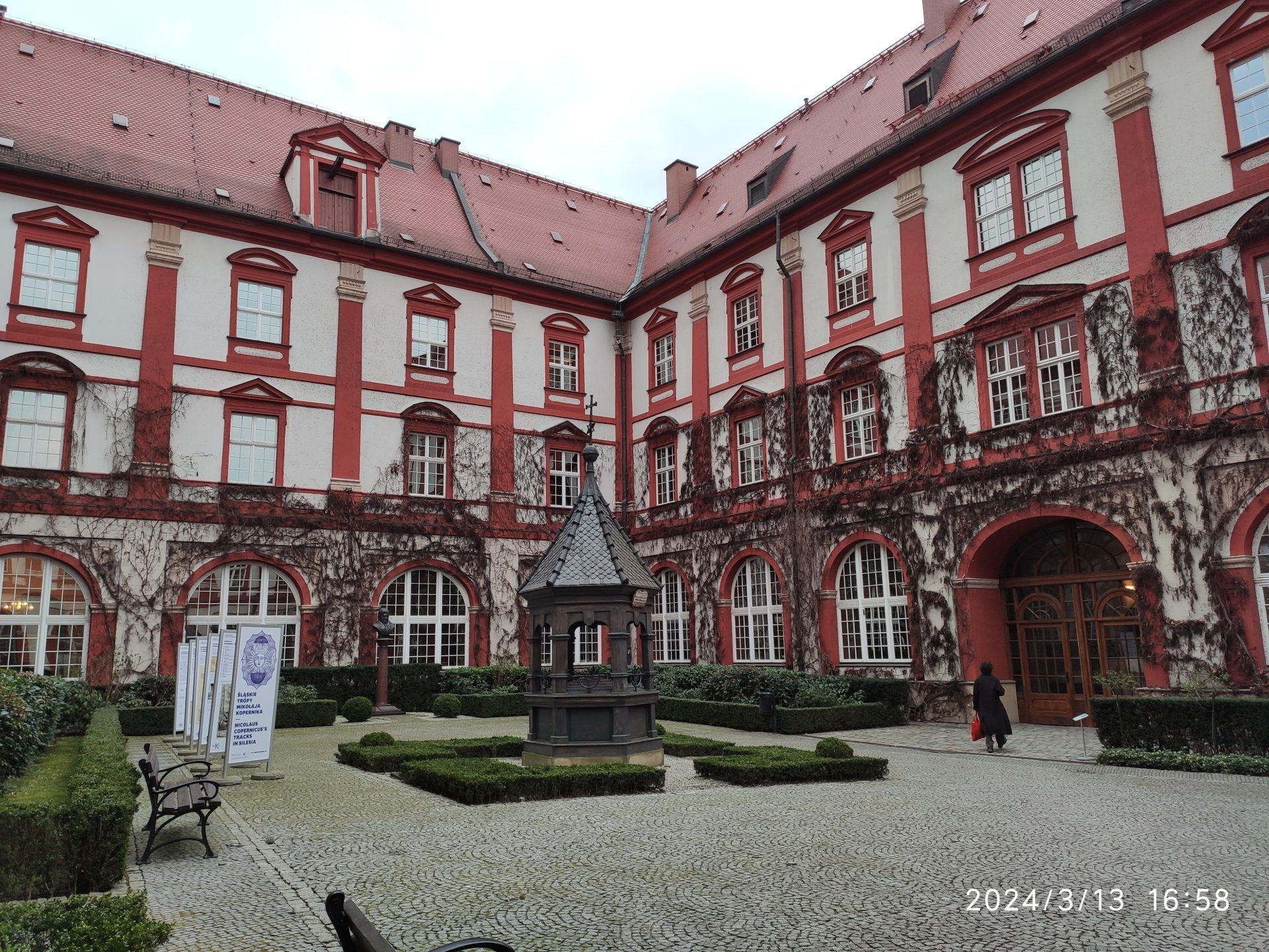 Dziedziniec zabytkowego budynku z czerwono-białą fasadą, ozdobioną suchym bluszczem i klasycznymi detalami architektonicznymi. Pośrodku znajduje się niewielka fontanna lub studnia, a całość otoczona jest przyciętymi krzewami i brukowaną nawierzchnią.