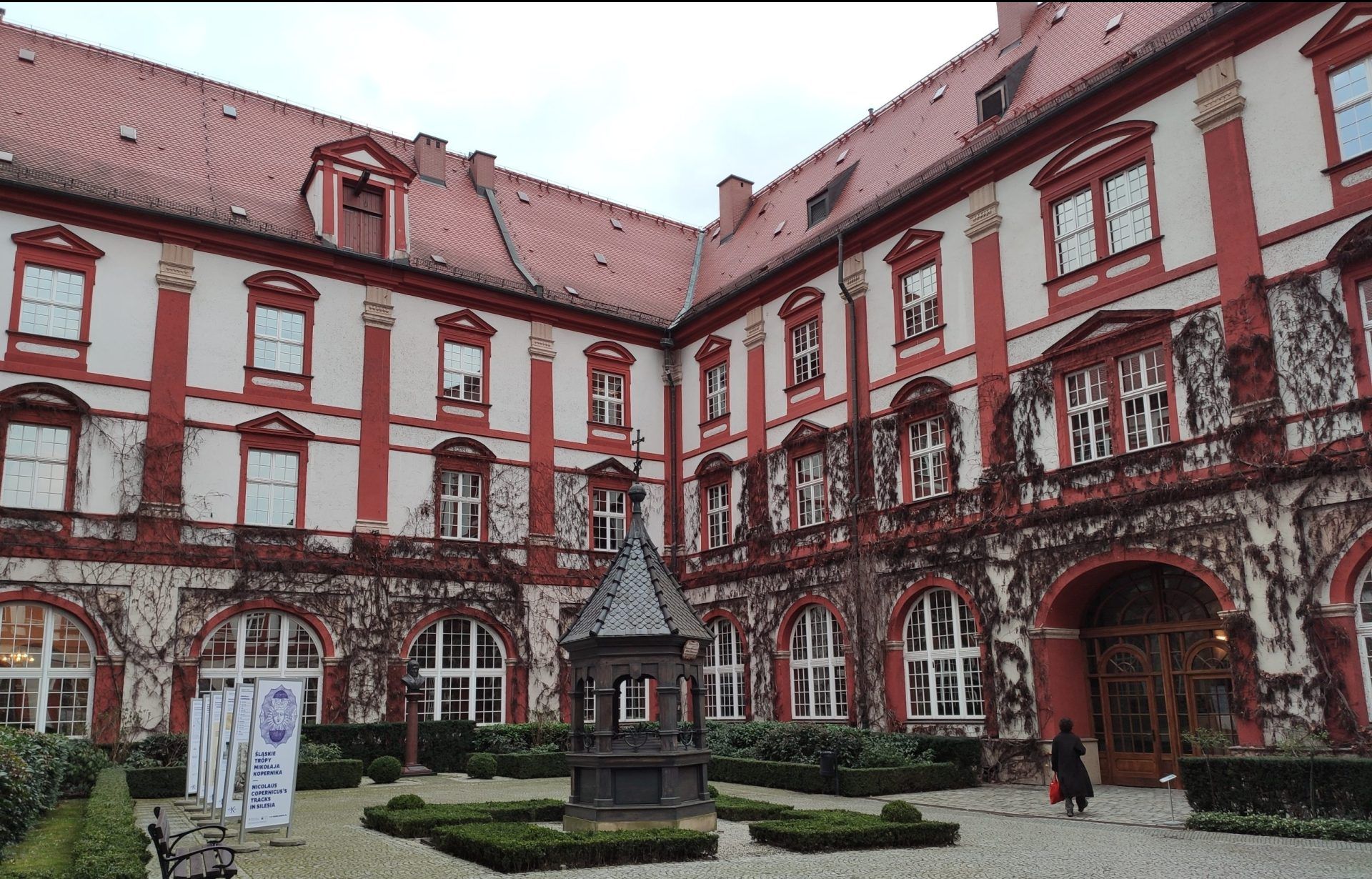 Dziedziniec zabytkowego budynku z czerwono-białą fasadą, ozdobioną suchym bluszczem i klasycznymi detalami architektonicznymi. Pośrodku znajduje się niewielka fontanna lub studnia, a całość otoczona jest przyciętymi krzewami i brukowaną nawierzchnią.