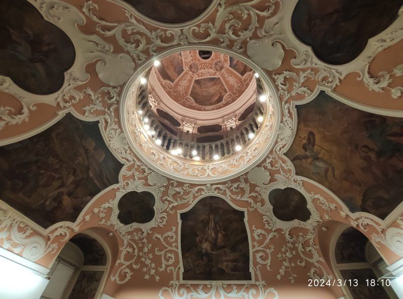 Bogato zdobiony sufit z barokowymi ornamentami i malowidłami przedstawiającymi sceny religijne lub mitologiczne. W centralnej części widać kopułę z balustradą i oświetleniem, która dodaje wnętrzu monumentalnego charakteru.