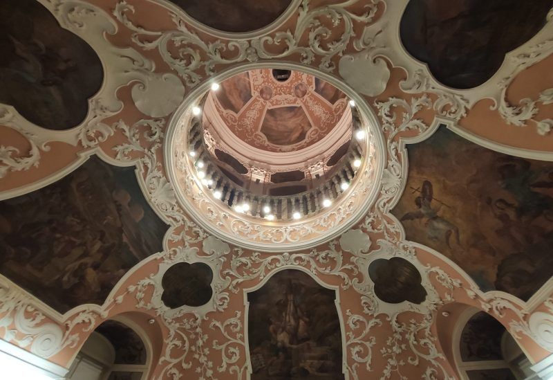 Bogato zdobiony sufit z barokowymi ornamentami i malowidłami przedstawiającymi sceny religijne lub mitologiczne. W centralnej części widać kopułę z balustradą i oświetleniem, która dodaje wnętrzu monumentalnego charakteru.