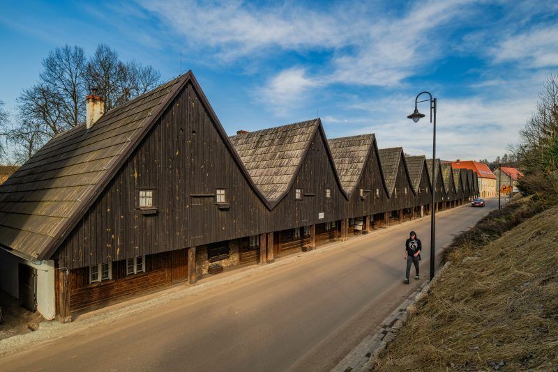 Drewniane domki Dwunastu Apostołów w Chełmsku Śląskim stoją w szeregu, z ciemnymi, szachulcowymi ścianami i spadzistymi dachami, otoczone drzewami, z błękitnym niebem w tle.