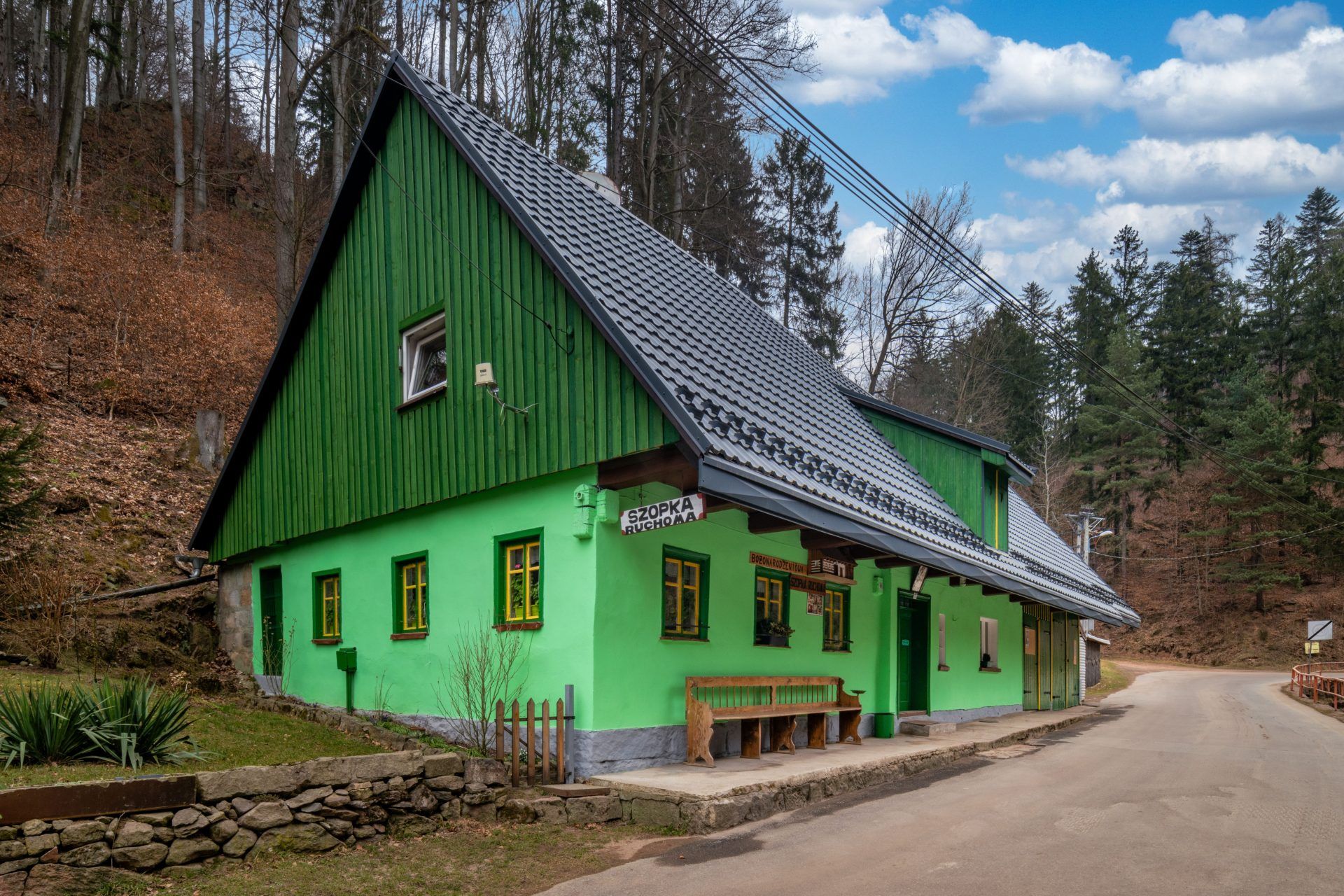 Na zdjęciu widać mały, zielony domek ze spadzistym dachem i żółtymi oknami, stojący wśród zieleni w spokojnym, wiejskim otoczeniu.