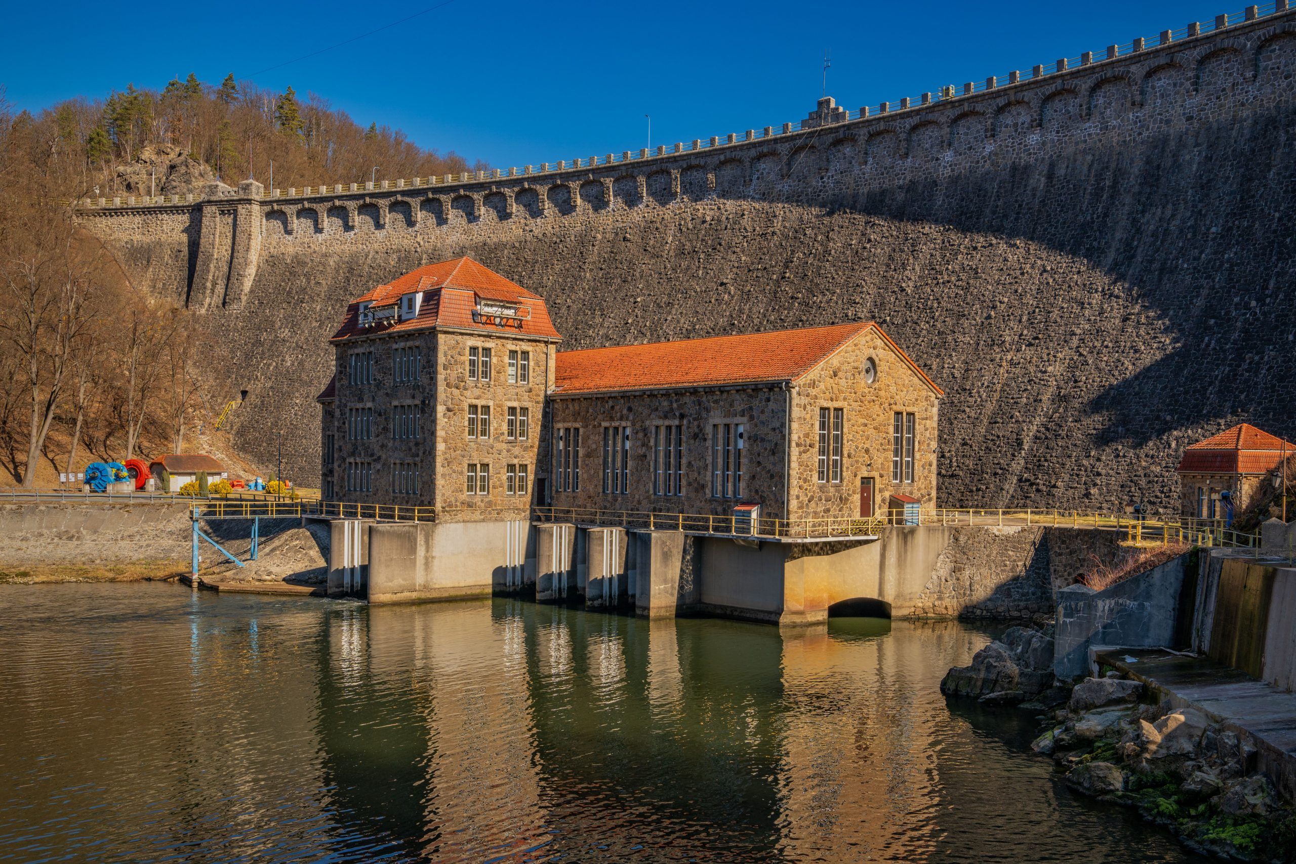 Na zdjęciu widać potężną kamienną zaporę w Pilchowicach, majestatycznie rozciągającą się wśród gór i odbijającą się w spokojnej tafli jeziora.