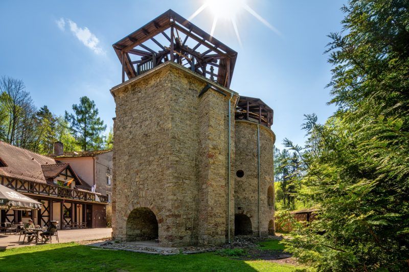 Solidny, prostokątny budynek z kamienia, z dwoma dużymi, drewnianymi kołami po bokach, otoczony gęstą, zieloną roślinnością i drzewami.