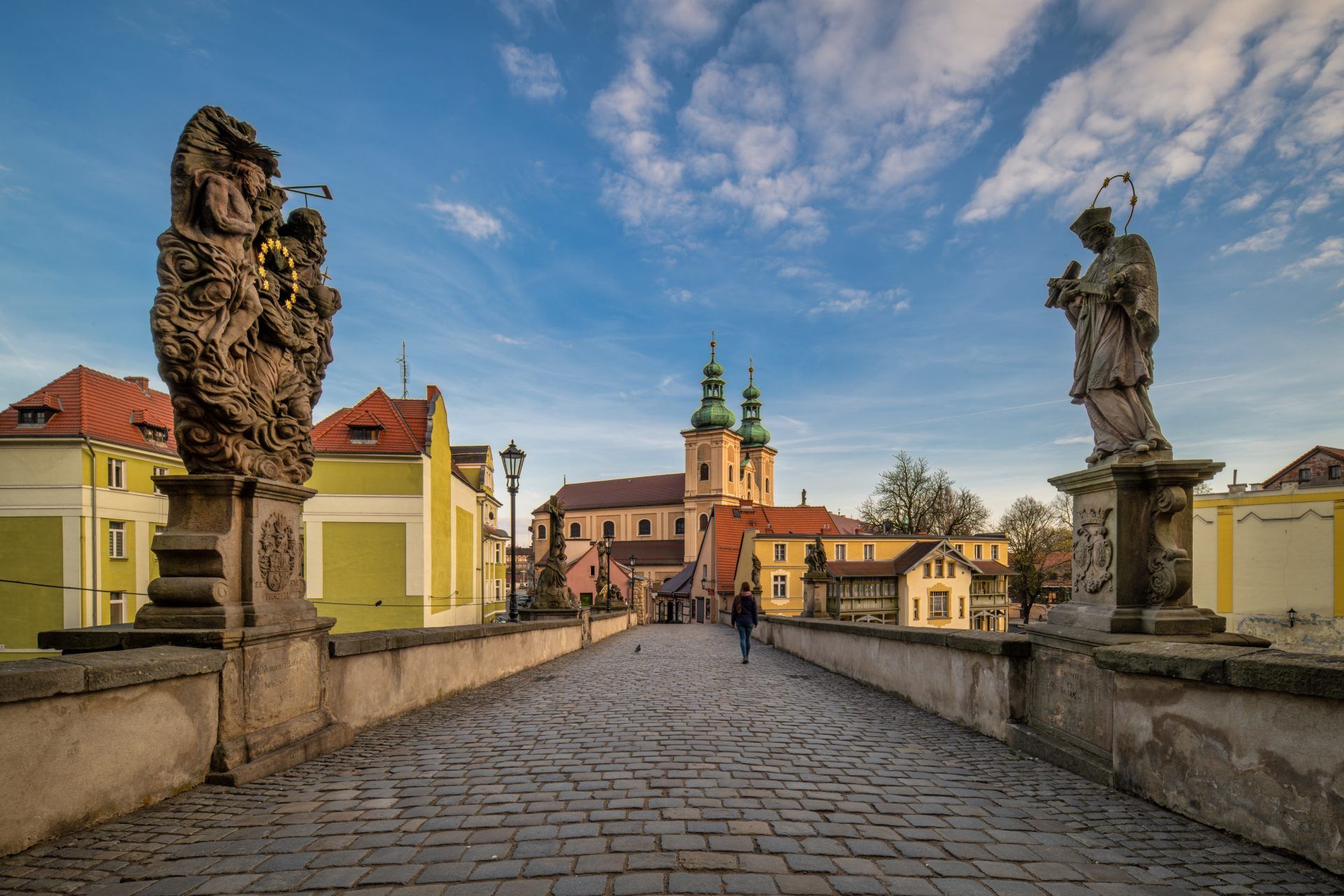 Na zdjęciu widać stary, kamienny most ozdobiony barokowymi rzeźbami świętyc, w tle z malowniczymi kamieniczkami kłodzkiej starówki.
