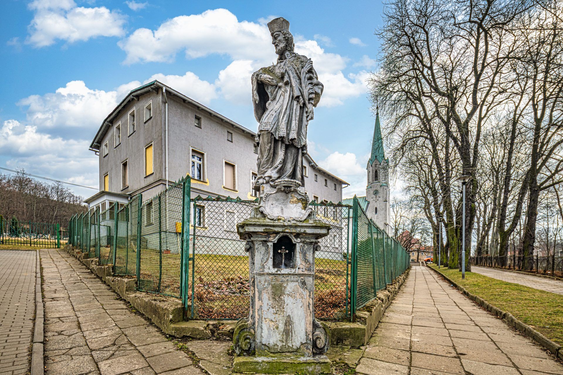 Na zdjęciu widać kamienną figurę św. Jana Nepomucena z krzyżem w dłoniach, stojącą na ozdobnym cokole.