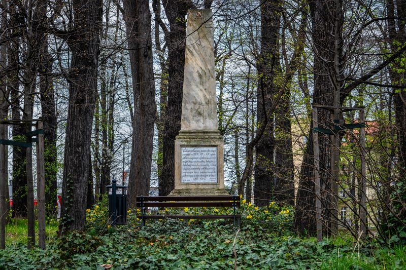 Kamienny obelisk stoi pośród zieleni parku, otoczony drzewami i spokojną alejką.