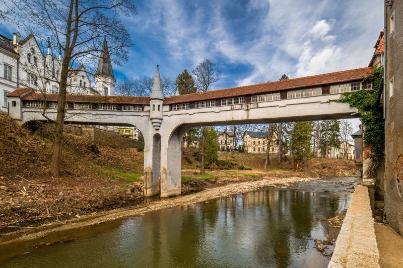 Kryty Most w Lądku-Zdroju to zabytkowa kamienna konstrukcja z łukowym przejściem, przykryta drewnianym dachem, otoczona bujną roślinnością i malowniczym krajobrazem.