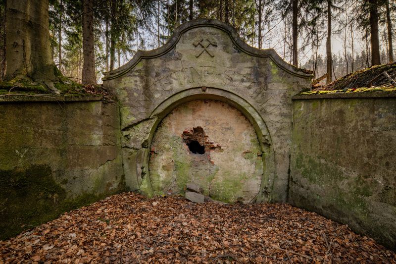 Na zdjęciu widać kamienny portal wejściowy sztolni Ernestine, otoczony przez zarośnięte wzgórze, z surowymi skałami i ścianą góry w tle.