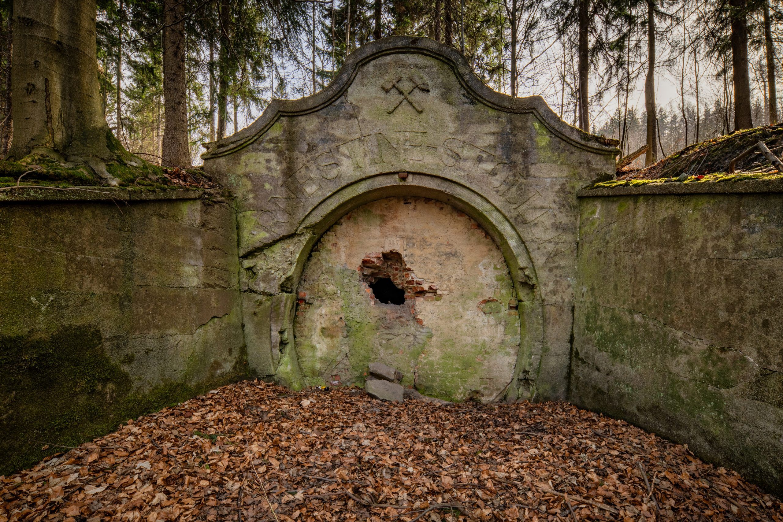Na zdjęciu widać kamienny portal wejściowy sztolni Ernestine, otoczony przez zarośnięte wzgórze, z surowymi skałami i ścianą góry w tle.