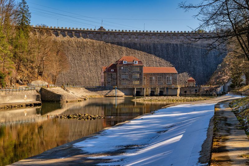 Na zdjęciu widać potężną kamienną zaporę w Pilchowicach, majestatycznie rozciągającą się wśród gór i odbijającą się w spokojnej tafli jeziora.