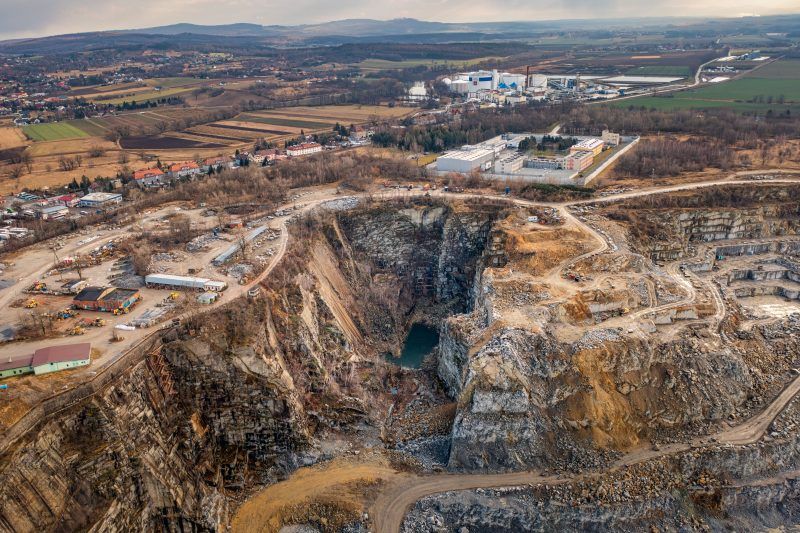 Na zdjęciu widać potężne, skalne ściany kamieniołomu w Strzelinie, z których wydobywa się granit, otoczone przez naturalną roślinność i malownicze jezioro w wykopach.