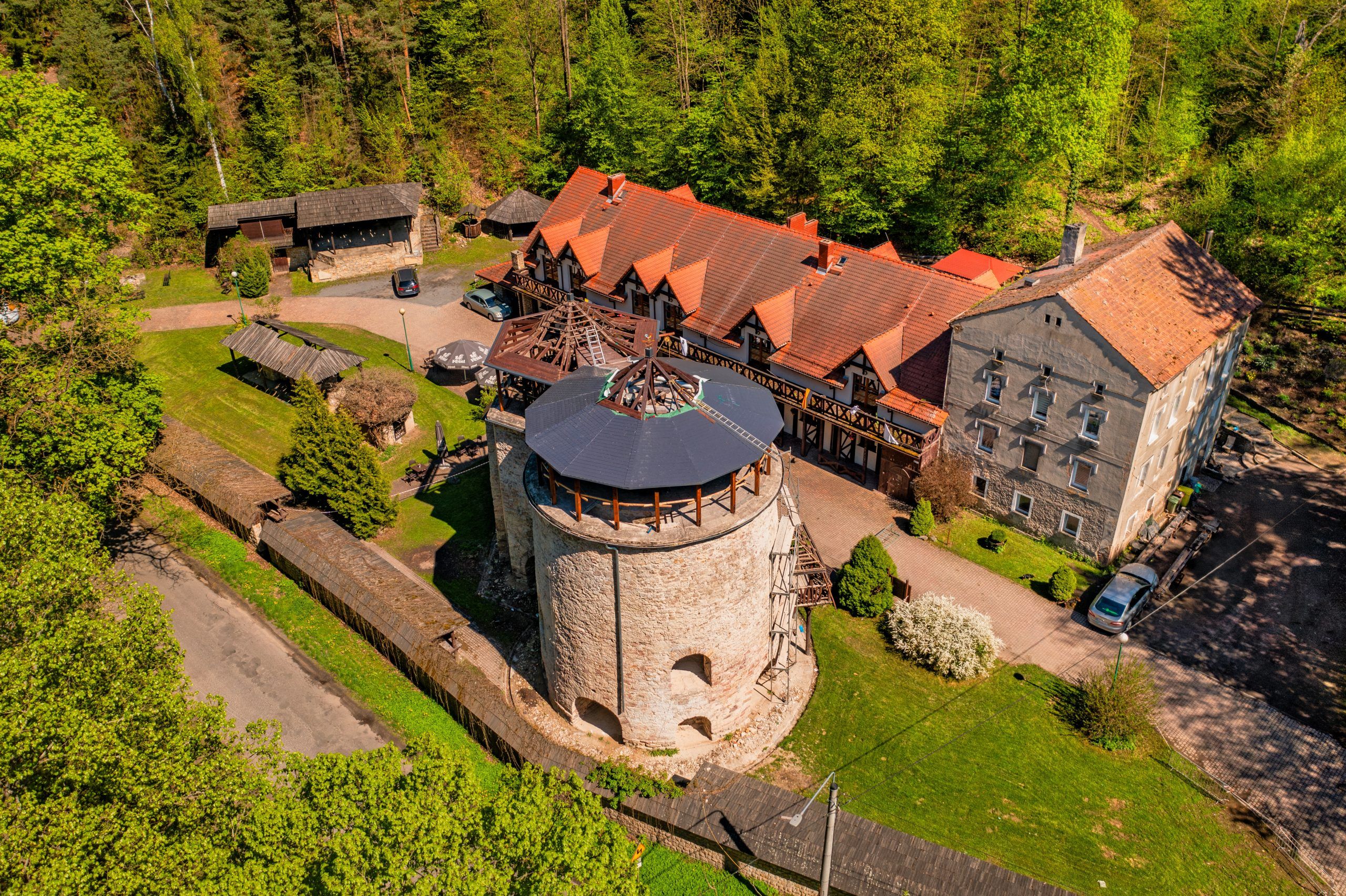 Z lotu ptaka widać solidny, prostokątny budynek z kamienia, z dwoma dużymi, drewnianymi kołami po bokach, otoczony gęstą, zieloną roślinnością i drzewami.
