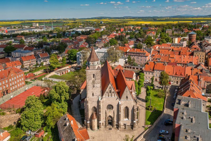 Monumentalna gotycka świątynia z wysoką wieżą i masywnymi, ceglastymi murami dominuje nad okoliczną zabudową.