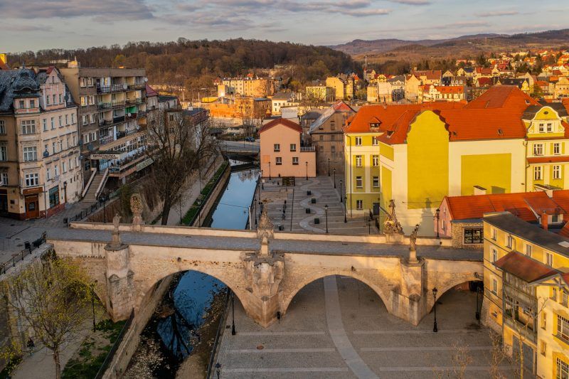 Na zdjęciu widać stary, kamienny most ozdobiony barokowymi rzeźbami świętych, rozciągający się łagodnym łukiem nad rzeką, w tle malownicze kamienice kłodzkiej starówki.