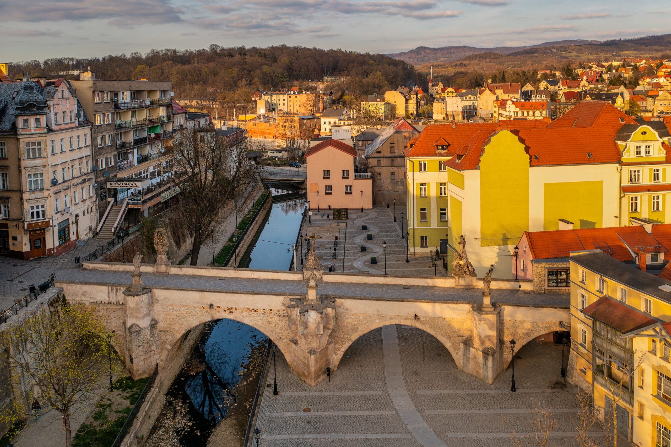 Na zdjęciu widać stary, kamienny most ozdobiony barokowymi rzeźbami świętych, rozciągający się łagodnym łukiem nad rzeką, w tle malownicze kamienice kłodzkiej starówki.