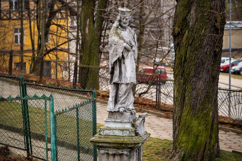 Na zdjęciu widać kamienną figurę św. Jana Nepomucena z krzyżem w dłoniach, stojącą na ozdobnym cokole.