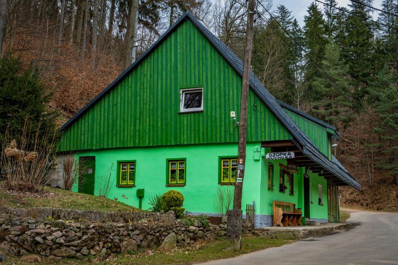 Na zdjęciu widać mały, zielony domek ze spadzistym dachem i żółtymi oknami, stojący wśród zieleni w spokojnym, wiejskim otoczeniu.