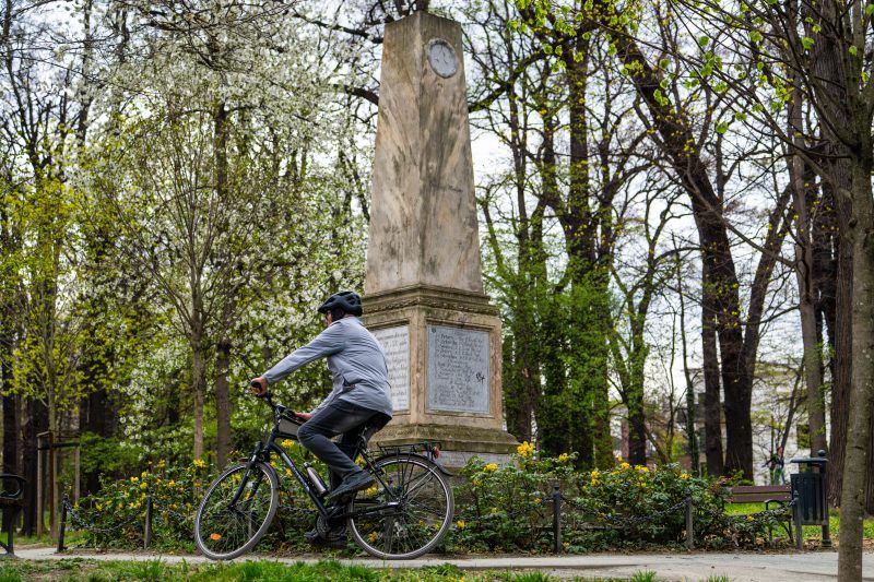 Kamienny obelisk stoi pośród zieleni parku, otoczony drzewami i spokojną alejką.