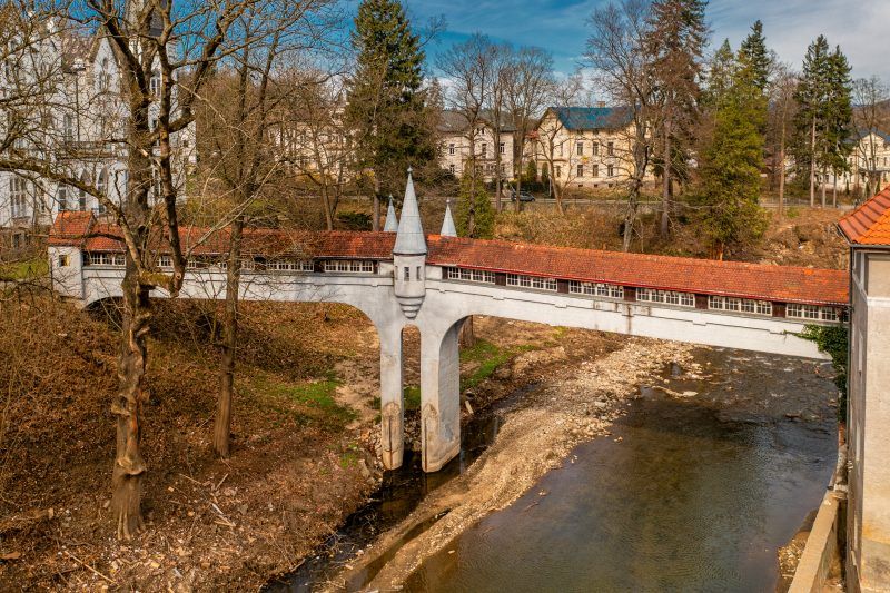 Kryty Most w Lądku-Zdroju to zabytkowa kamienna konstrukcja z łukowym przejściem, przykryta drewnianym dachem, otoczona bujną roślinnością i malowniczym krajobrazem.