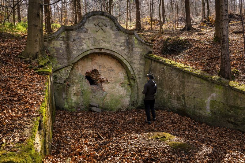 Na zdjęciu widać kamienny portal wejściowy sztolni Ernestine, otoczony przez zarośnięte wzgórze, z surowymi skałami i ścianą góry w tle.