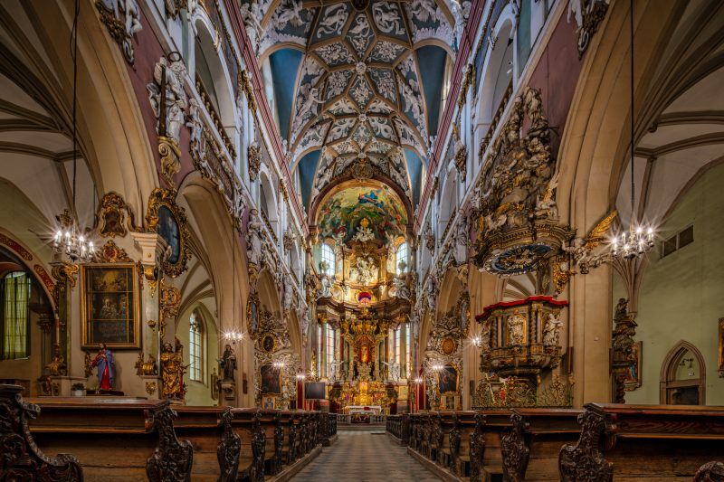 Wnętrze kościoła wypełniają wysokie, gotyckie sklepienia, smukłe filary i ciepłe światło przenikające przez witraże nad bogato zdobionymi ołtarzami.