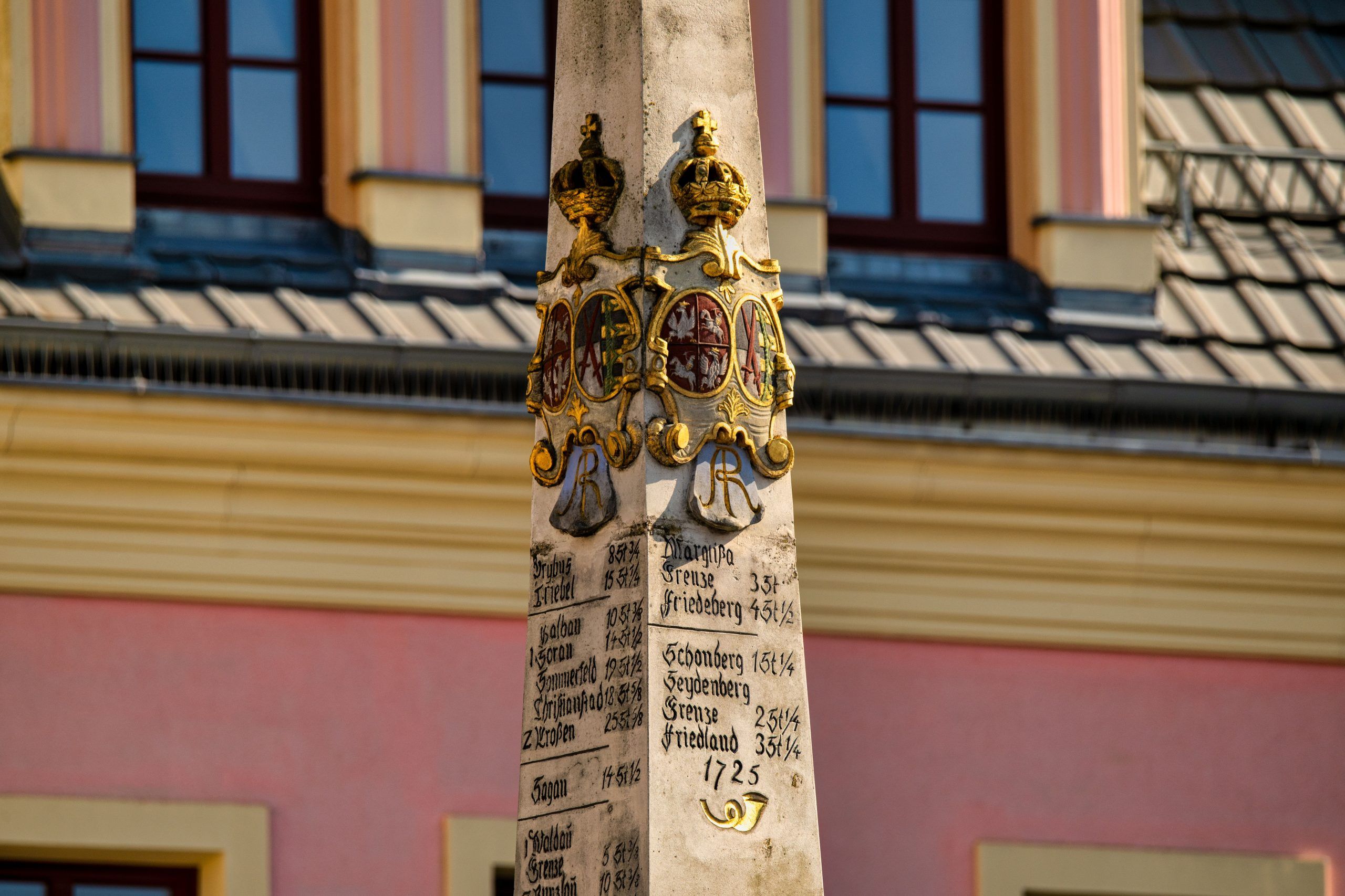 Kamienny, barokowy słup z herbami i inskrypcjami wskazującymi odległości do miast, w tle różowy budynek.