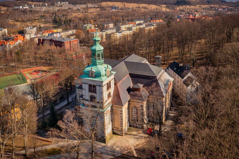 Na zdjęciu widać okazały Kościół Łaski w Kamiennej Górze – barokową budowlę o jasnej elewacji, wysokim dachu i smukłej wieży górującej nad otaczającym ją parkiem.