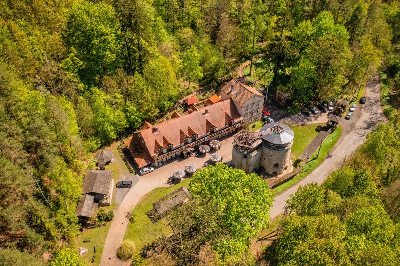 Z lotu ptaka widać solidny, prostokątny budynek z kamienia, z dwoma dużymi, drewnianymi kołami po bokach, otoczony gęstą, zieloną roślinnością i drzewami.