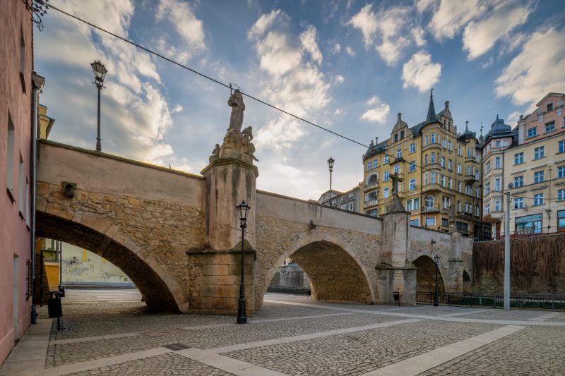 Na zdjęciu widać stary, kamienny most ozdobiony barokowymi rzeźbami świętych, rozciągający się łagodnym łukiem, w tle malownicze kamieniczki kłodzkiej starówki.