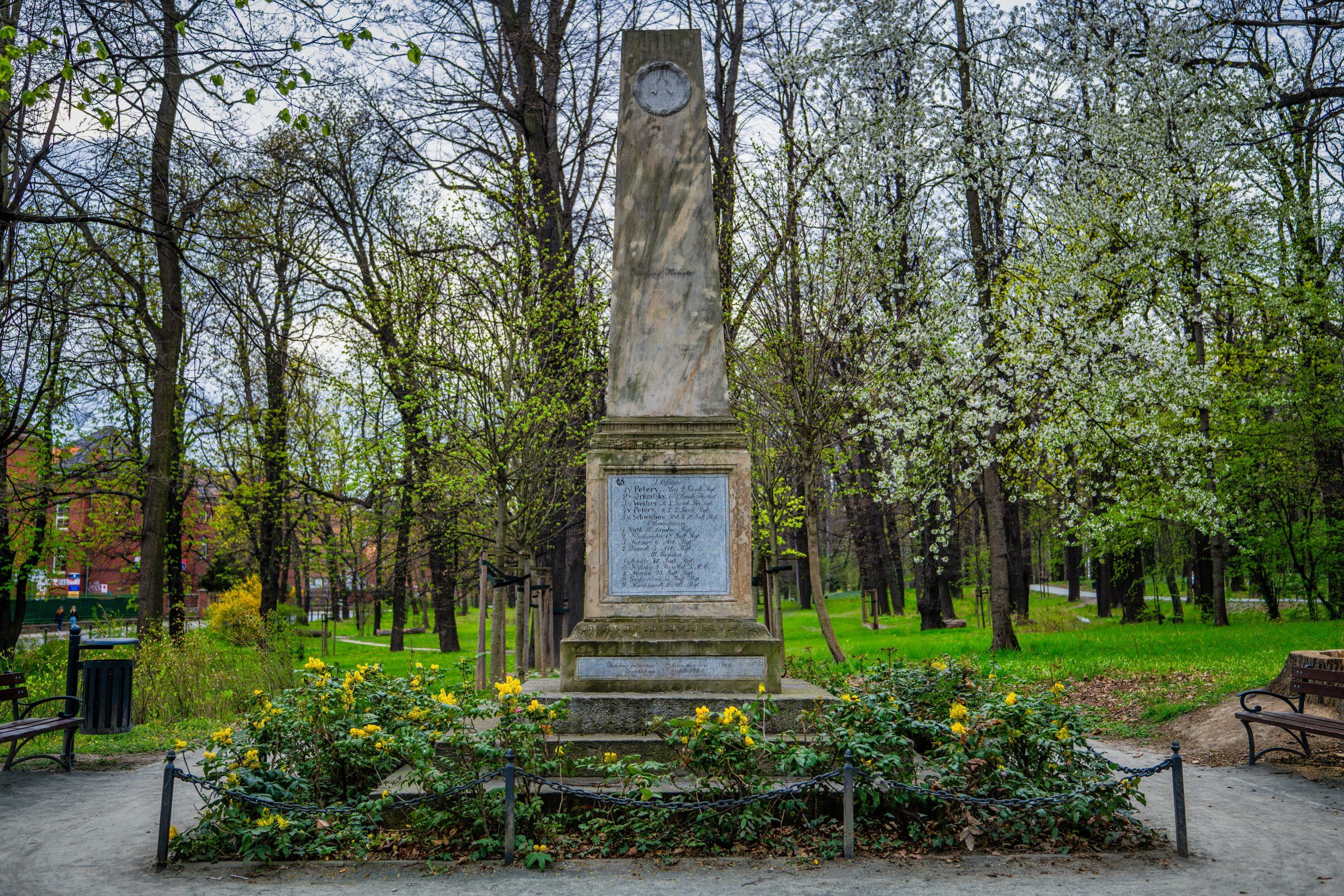 Kamienny obelisk stoi pośród zieleni parku, otoczony drzewami i spokojną alejką.