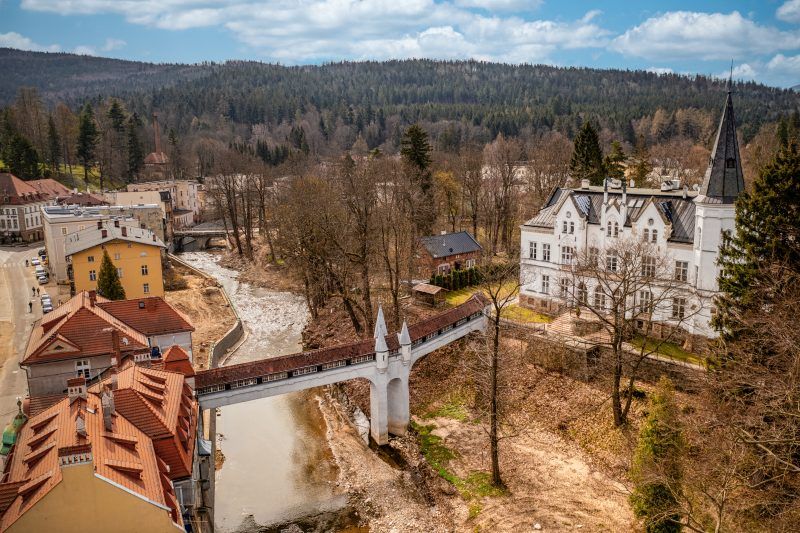 Kryty Most w Lądku-Zdroju to zabytkowa kamienna konstrukcja z łukowym przejściem, przykryta drewnianym dachem, otoczona bujną roślinnością i malowniczym krajobrazem.