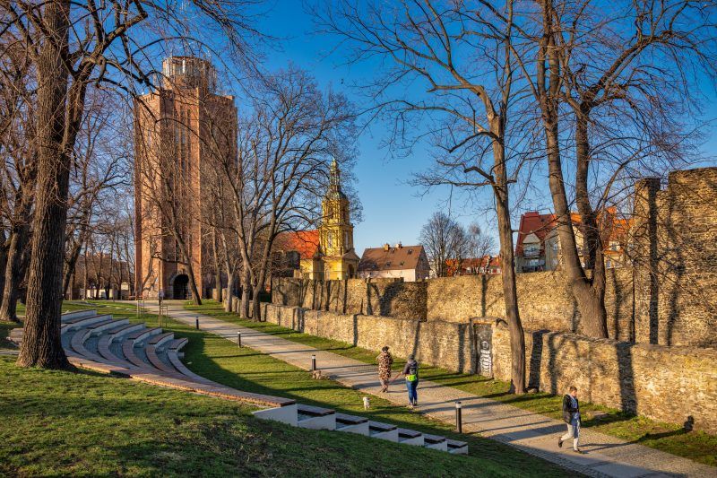 Na zdjęciu widać fragment masywnych, szarych kamiennych murów obronnych z prostokątnymi otworami strzelniczymi i częściowo zachowaną basztą, otoczonych trawnikiem i zielenią.