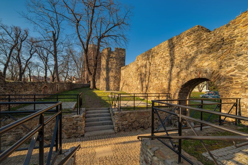 Na zdjęciu widać fragment masywnych, szarych kamiennych murów obronnych z prostokątnymi otworami strzelniczymi i częściowo zachowaną basztą, otoczonych trawnikiem i zielenią.