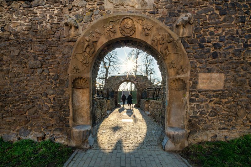 Na zdjęciu widać fragment masywnych, szarych kamiennych murów obronnych z prostokątnymi otworami strzelniczymi i częściowo zachowaną basztą, otoczonych trawnikiem i zielenią.
