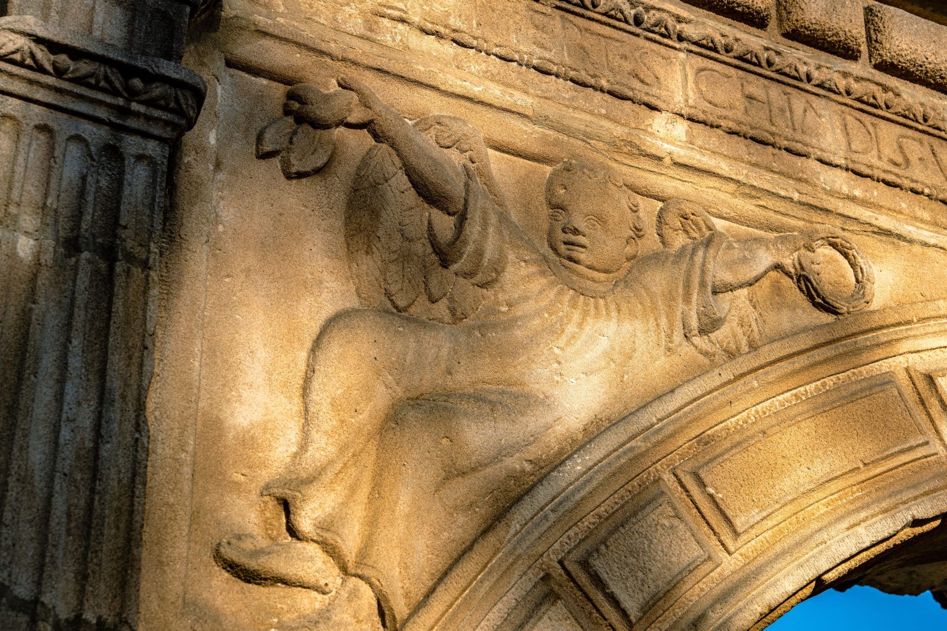 Na zbliżeniu widać wyryty w kamieniu drobny wizerunek aniołka z rozłożonymi skrzydłami, o delikatnie zaznaczonych rysach twarzy i subtelnych detalach szat, kontrastujący z surową powierzchnią muru.