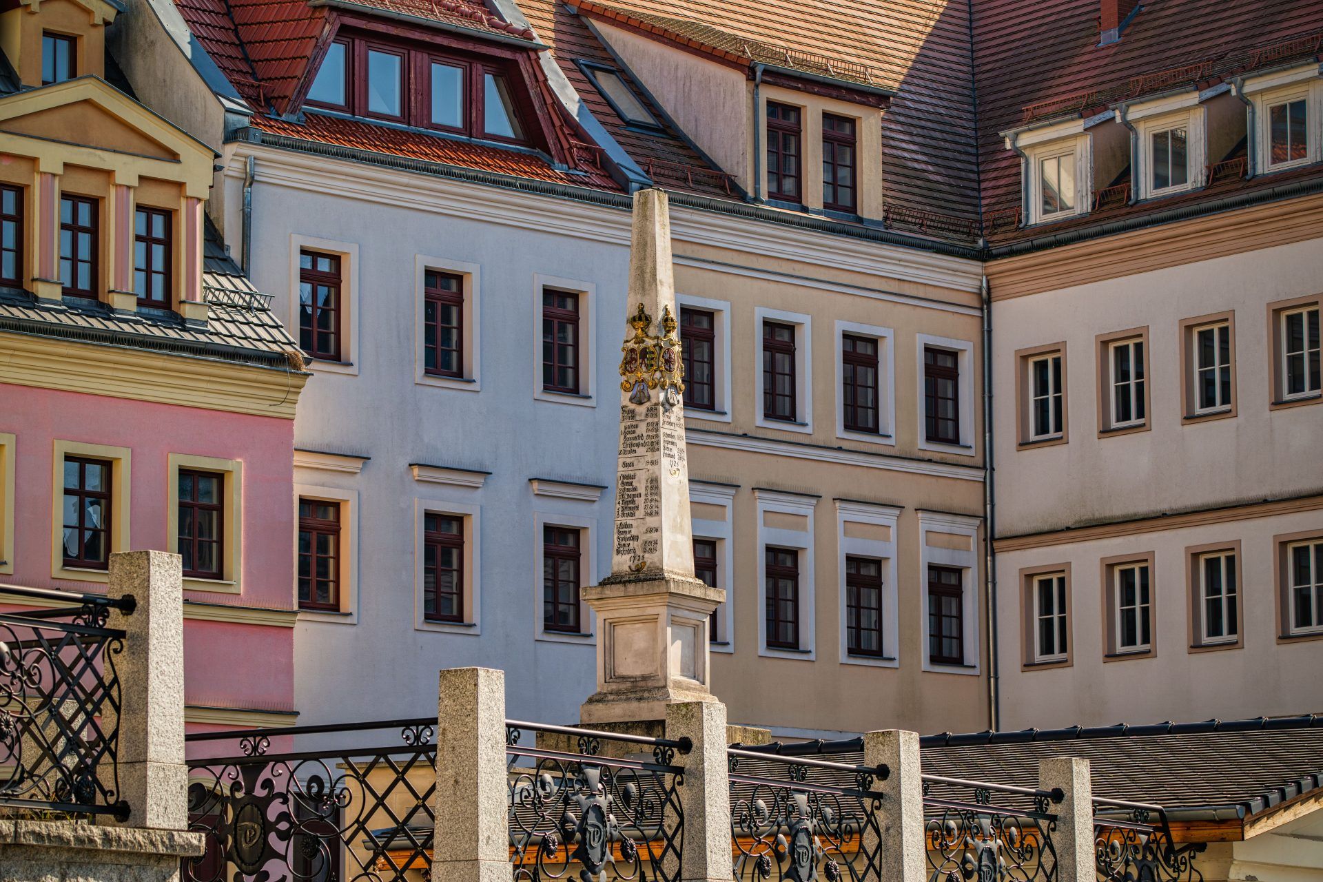 Kamienny, barokowy słup z herbami i inskrypcjami wskazującymi odległości do miast, w tle jasne budynki.