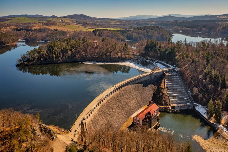 Na zdjęciu widać potężną kamienną zaporę w Pilchowicach, majestatycznie rozciągającą się wśród gór i odbijającą się w spokojnej tafli jeziora.