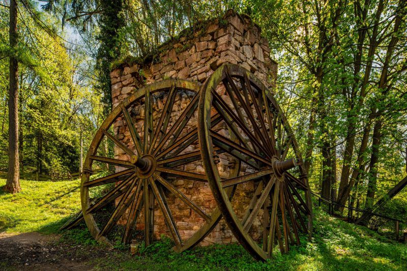 Zdjęcie pokazuje fragment starej, kamiennej konstrukcji z dwoma dużymi, drewnianymi kołami osadzonymi po bokach, otoczonej zielenią lasu.