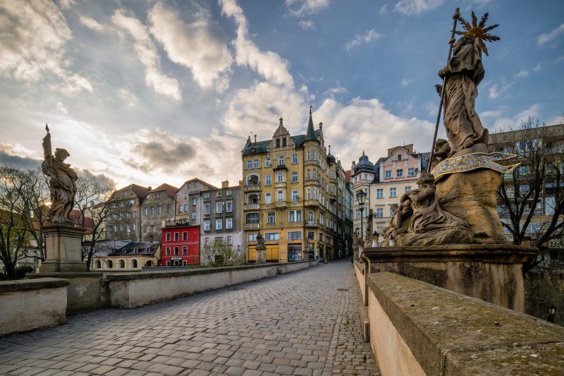 Na zdjęciu widać stary, kamienny most ozdobiony barokowymi rzeźbami świętych, w tle malownicze kamieniczki kłodzkiej starówki.