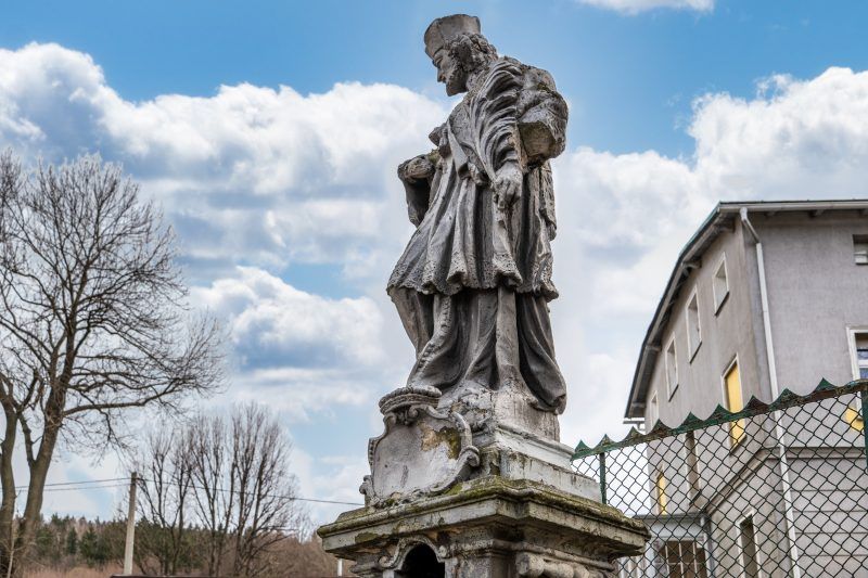 Na zdjęciu widać kamienną figurę św. Jana Nepomucena z krzyżem w dłoniach, stojącą na ozdobnym cokole.