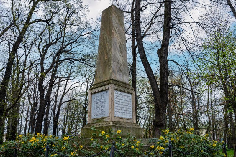 Kamienny obelisk stoi pośród zieleni parku, otoczony drzewami i spokojną alejką.