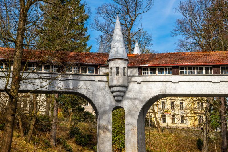 Kryty Most w Lądku-Zdroju to zabytkowa kamienna konstrukcja z łukowym przejściem, przykryta drewnianym dachem, otoczona bujną roślinnością i malowniczym krajobrazem.