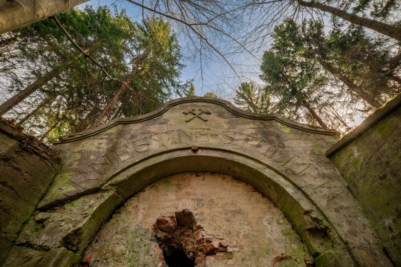 Na zdjęciu widać kamienny portal wejściowy sztolni Ernestine, otoczony przez zarośnięte wzgórze, z surowymi skałami i ścianą góry w tle.