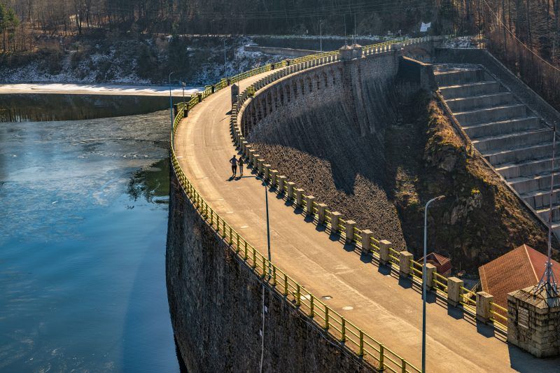 Na zdjęciu widać potężną kamienną zaporę w Pilchowicach, majestatycznie rozciągającą się wśród gór i odbijającą się w spokojnej tafli jeziora.