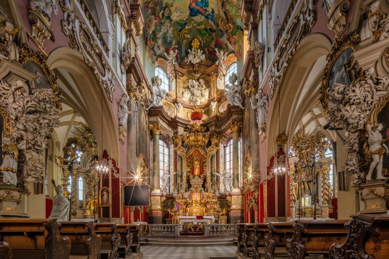 Wnętrze kościoła wypełniają wysokie, gotyckie sklepienia, smukłe filary i ciepłe światło przenikające przez witraże nad bogato zdobionymi ołtarzami.