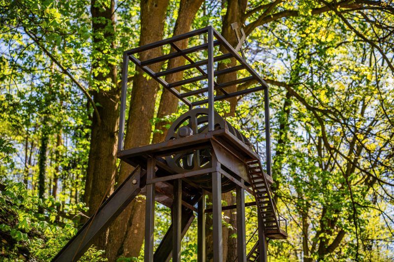 Zdjęcie przedstawia metalową konstrukcję szybu górniczego na tle bujnej zieleni drzew w słoneczny dzień.