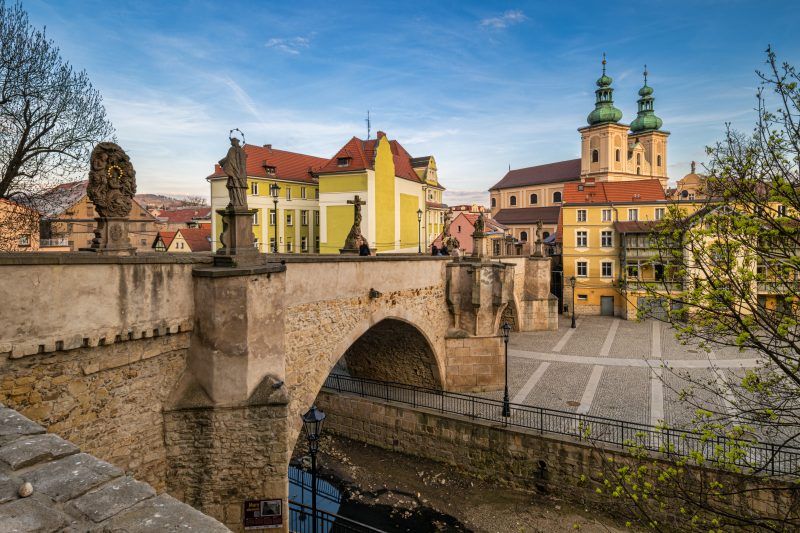 Na zdjęciu widać stary, kamienny most ozdobiony barokowymi rzeźbami świętych, rozciągający się łagodnym łukiem nad rzeką, z tle malowniczymi kamieniczkami kłodzkiej starówki.